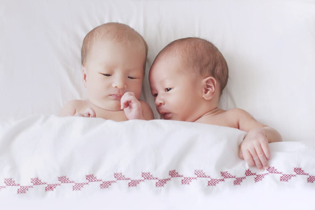 Dois bebês dormindo juntos na cama, com uma expressão tranquila, aconchegados sob um cobertor branco com detalhes vermelhos, criando uma imagem de fofura e conexão.
