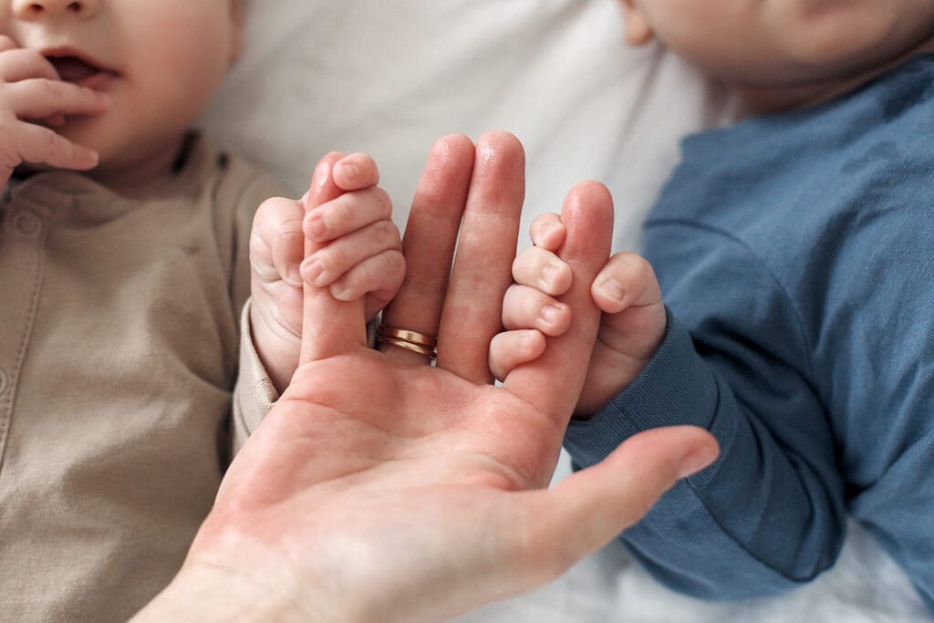 Imagem tocando a mão de duas crianças pequenas com um adulto, simbolizando cuidado e amor familiar, com foco na delicadeza do momento.